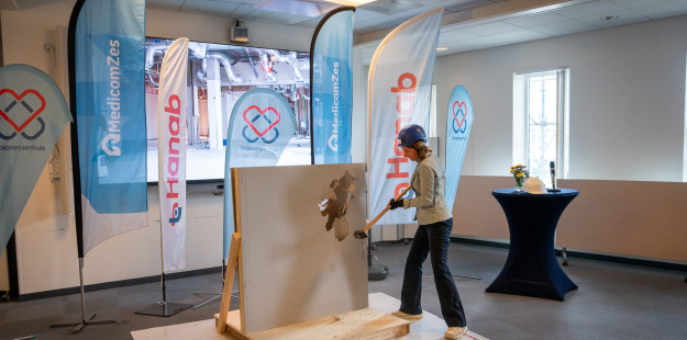 Een persoon met een veiligheidshelm en veiligheidsbril slaat met een grote hamer door een wandpaneel. De setting is een binnenruimte met banners van MedicomZes en Hanab op de achtergrond. De actie symboliseert waarschijnlijk de officiële start of een symbolische doorbraak van het nieuwbouwproject.