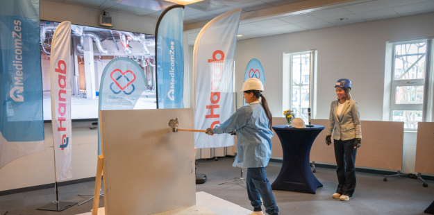 Een andere persoon met beschermende uitrusting (helm en bril) slaat met een hamer op een wandpaneel, terwijl een collega toekijkt. De banners van MedicomZes en Hanab zijn opnieuw aanwezig. Deze foto toont de actieve deelname aan het ceremoniële moment van de doorbraak.