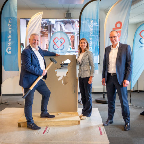 Drie personen in nette kleding staan rondom het doorbroken wandpaneel. Eén van hen houdt een hamer vast, terwijl de anderen toekijken. Ook hier zijn de banners van MedicomZes en Hanab zichtbaar. Deze foto benadrukt de betrokkenheid van belangrijke vertegenwoordigers bij het evenement.