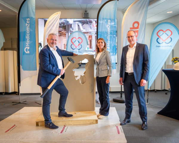 Drie personen in nette kleding staan rondom het doorbroken wandpaneel. Eén van hen houdt een hamer vast, terwijl de anderen toekijken. Ook hier zijn de banners van MedicomZes en Hanab zichtbaar. Deze foto benadrukt de betrokkenheid van belangrijke vertegenwoordigers bij het evenement.