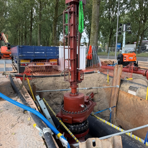 Werkzaamheden aan een ondergrondse leiding, met centraal een grote roodbruine leidingconstructie in een open bouwput. Rondom liggen leidingen, slangen en bouwmaterialen en op de achtergrond staan bouwmaterieel, afzettingen en bomen.