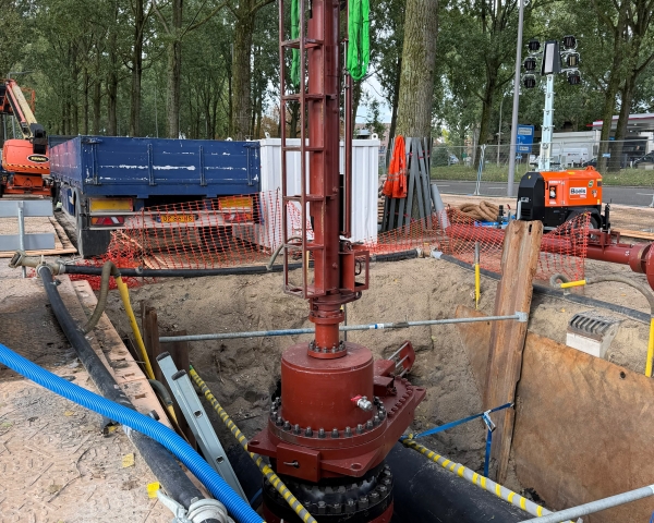 Werkzaamheden aan een ondergrondse leiding, met centraal een grote roodbruine leidingconstructie in een open bouwput. Rondom liggen leidingen, slangen en bouwmaterialen en op de achtergrond staan bouwmaterieel, afzettingen en bomen.